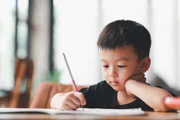 Asian Boy Doing homework with the intention. Child boy holding pencil writing, A boy drawing on white paper at the table, Elementary school and home schooling, Distance Education concept..