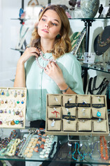 Fototapeta na wymiar Interested girl behind glass of showcase trying on elegant necklace from natural pink aventurine