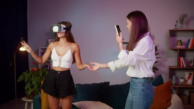 Young woman wearing virtual reality glasses, girlfriend recording video on smartphone. Sisters playing vr video game. Students enjoying AR 3D education learning video games in living room. 