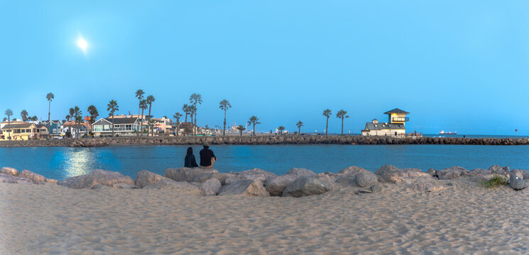 Couple enjoying the moon's reflection on the ocean sitting on the beach looking over at rocky pier with a white building  beacon and container ship in background at night time