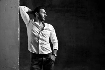 Black and white. Modern bearded, unshaved man in white shirt and jeans stands, leaning on concrete...
