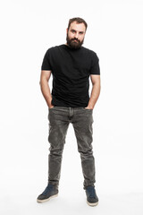 Fototapeta premium A man with a beard is standing with his hands in his pockets. Young man in jeans and a black T-shirt. Full height. White background. Vertical.