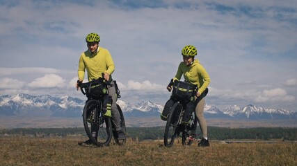 Fototapeta na wymiar The man and woman travel on mixed terrain cycle touring with bikepacking. The two people journey with bicycle bags. Sport bikepacking, bike, sportswear in green black colors. Mountain snow capped.