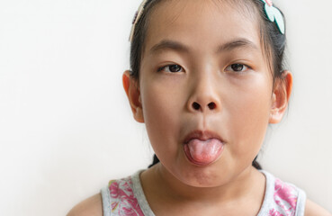 Headshot of Asian cute child girl with a grimacing face, black long hair tied, looks and shows tongue out at the camera, stands over white wall background, close up portrait.