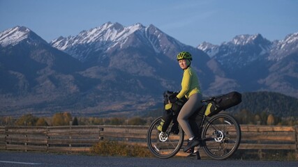 Fototapeta premium The woman travel on mixed terrain cycle touring with bikepacking. The traveler journey with bicycle bags. Sport tourism bikepacking, bike, sportswear in green black colors. Mountain snow capped.