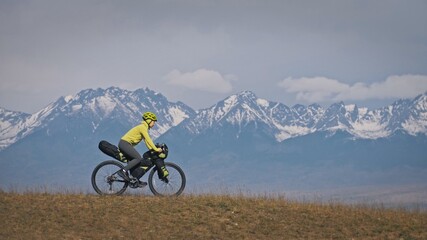 The woman travel on mixed terrain cycle touring with bikepacking. The traveler journey with bicycle bags. Sport tourism bikepacking, bike, sportswear in green black colors. Mountain snow capped.