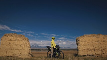 The woman travel on mixed terrain cycle touring with bikepacking. The traveler journey with bicycle bags. Sport bikepacking, bike, sportswear in green black colors. Mountain snow capped, stone arch.