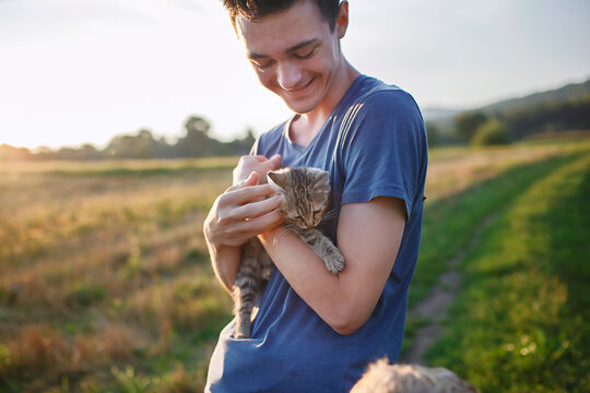 Young guy found and picked up little stripped kitten during walking in the field and going to home it, homeless domestic animals care, concept of volunteering and animal rescue
