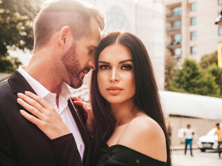 Beautiful fashion woman and her handsome elegant boyfriend in suit. Sexy brunette model in black evening dress. Fashionable couple posing in the street in Europe. Brutal man and his female outdoors