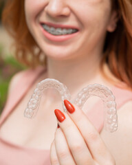 Beautiful young red-haired woman with braces on her teeth holding removable retainers outdoors