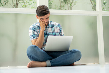 serious young man sitting on floor working with laptop computer on window at home . Bored worker tired from work at home office distracted from boring job