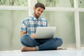 serious young man sitting on floor working with laptop computer on window at home . Bored worker tired from work at home office distracted from boring job