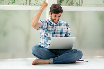 happy young man sitting on floor working with laptop computer raise  fist celebrating to succes  on window at home  . excited male hand up to win