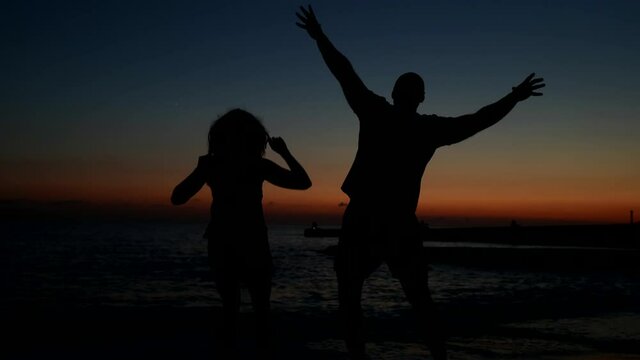 A happy couple jumps on the beach at night, enjoying the sunset. Conceptual video of happiness and bliss with people jumping for joy, celebrating in silhouette
