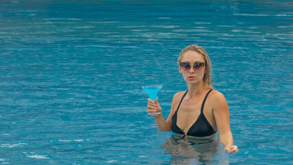 Middle shot pretty woman drinking blue cocktail alcohol liquor in swimming pool at hotel. Portrait of sexy girl in pool outdoor. Beautiful caucasian women with blond hair, black swimsuit, sunglasses.