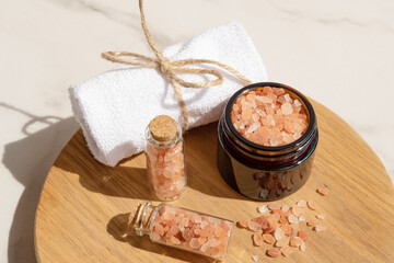 Pink Himalayan salt in the glass bottles with rolled towel on wooden board. Close up of pink crystals of sea salt. Spa body care concept