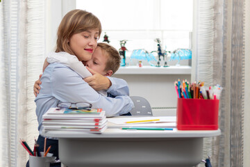 A beautiful young mother with her son is doing homework at a desk at home against a light background of a window during a pandemic quarantine. Selective focus. Portrait