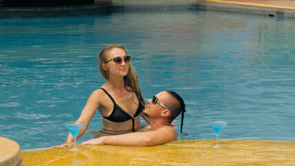 The loving couple hugs and kisses, drinking blue cocktail alcohol liquor in swimming pool at hotel...