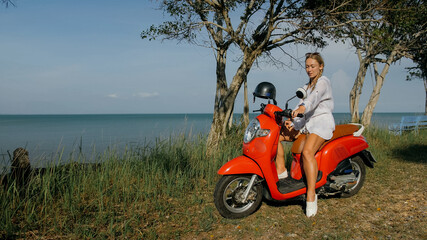 Fototapeta premium Woman on red motorbike in white clothes drive on forest road trail trip. One girl caucasian tourist go on scooter, nearby tropical palm tree. Asia Thailand ride tourism. Motorcycle rent Safety helmet.