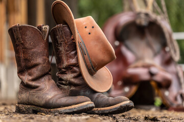 Ranch life scenery: muddy western boots in front of a western saddle. Cowboy boots. Muddy working boots - obrazy, fototapety, plakaty