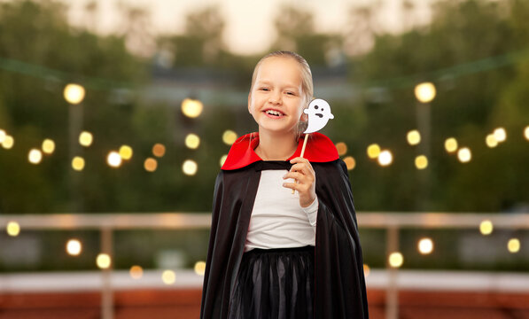 halloween, holiday and childhood concept - happy smiling girl in black dracula costume with cape and ghost over garland lights at roof top party background