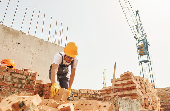 Using bricks. Young construction worker in uniform is busy at the unfinished building