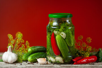 Pickled cucumbers in glass jars and spices and vegetables for preparation of pickles on red background