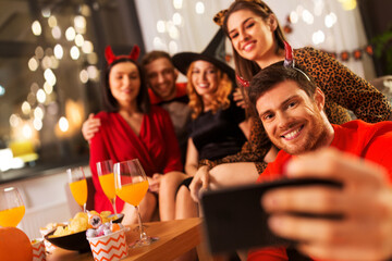 friendship, holiday and people concept - group of happy smiling friends in halloween costumes of vampire, devil, witch and cheetah taking selfie by smartphone at home party at night