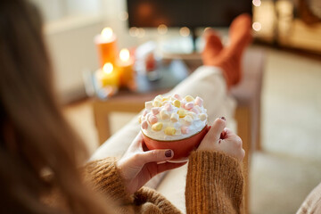 halloween, holidays and leisure concept - close up of young woman watching tv and holding mug of marshmallow and whipped cream with her feet on table at cozy home