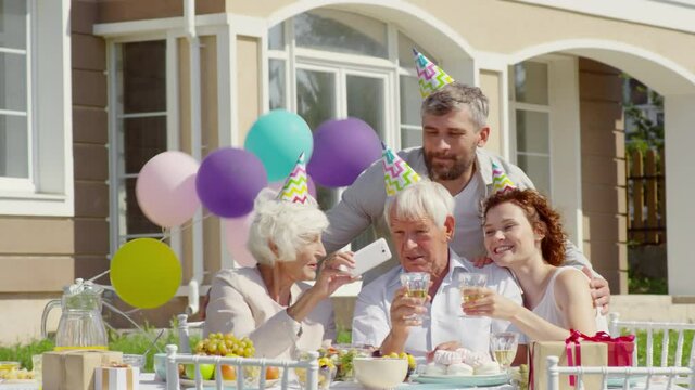 Happy grandparents and young family couple in party hats sitting at dinner table outdoors, smiling and posing with wine glasses while taking a selfie with smartphone on birthday celebration