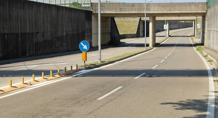 A four-lane road, with barrier on the middle, passes under a  gray concrete underpass and alongside a wall of the same color. Concrete sidewalk and traffic signs. Background for copy space - obrazy, fototapety, plakaty
