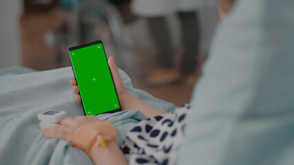 Sick girl holding mock up green screen chroma key phone with isolated display during recovery consultation in hospital ward. Child with oximeter on finger resting in bed after suffering medical
