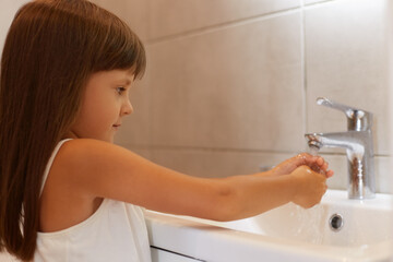Side view portrait of cute female child with dark hair wearing white sleeveless t shirt, posing in bathroom near sink and washing her hands before going to bed.
