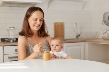 Obraz na płótnie Canvas Indoor shot of smiling female sitting at table in kitchen with baby girl in hands and feeding daughter with fruit or vegetable puree, complementary feeding of a child.