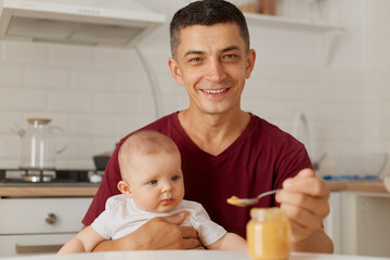 Obraz na płótnie Canvas Indoor portrait of happy young father wearing maroon t shirt holding baby on his knees and feeding son or daughter with vegetable or fruit puree in kitchen.