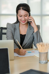 Young attractive Asian woman in grey business suit sitting talking on mobile phone in modern looking office with blurry windows background. Concept for modern office lifestyle