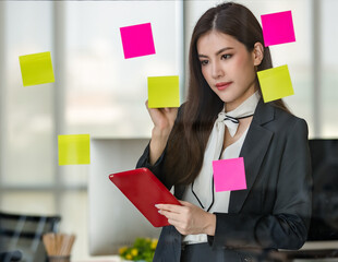 Young attractive Asian woman in black business writing on post it on glass panel one hand holding tablet in modern looking office with blurry windows background. Concept for modern office lifestyle