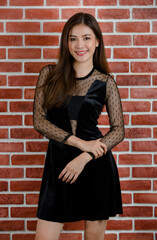 Portrait of young beautiful attractive Asian female model in black revealing dress smiling and standing against orange brick wall background