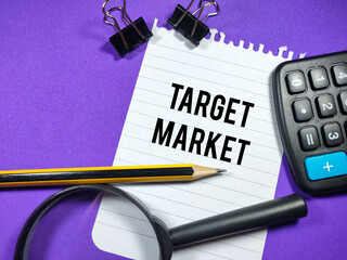 Business concept.Text TARGET MARKET writing on notepaper with magnifying glass,calculator,paper clips and pencil on purple background.
