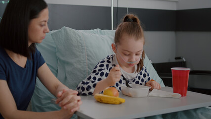 Mother sitting with sick daughter while eating healthy food meal in hospital ward during medical...