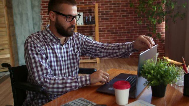 Confident hipster man in wheelchair finishes working on laptop, closes it and drives away. Concept of freelancing