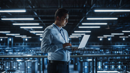 Portrait of IT Specialist Uses Laptop in Data Center. Server Farm Cloud Computing Facility with...