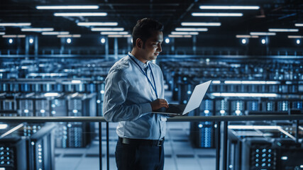 Portrait of IT Specialist Uses Laptop in Data Center. Server Farm Cloud Computing Facility with Male Maintenance Administrator Working. Cyber Security and Network Protection.