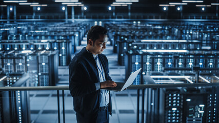 Portrait of IT Specialist Uses Laptop in Data Center. Server Farm Cloud Computing Facility with...