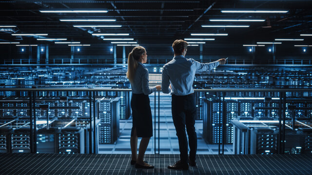 Data Center Male It Specialist and Female System administrator Talk, Use Laptop. Server Farm Cloud Computing Facility with Two Information Technology Professionals working on Cyber Secury.