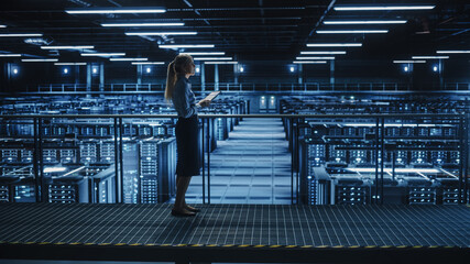 Female IT Specialist Using Tablet Computer in Data Center, Walking on a Bridge Overlooking Gigantic Server Farm Cloud Computing Facility. Succesful Businesswoman, e-Business Entrepreneur.