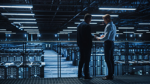 Data Center IT Specialist and e-Business Enrepreneur Talk, Use Laptop. Two Information Technology Professionals Walking on Bridge Overlooking Cloud Computing Server Farm.