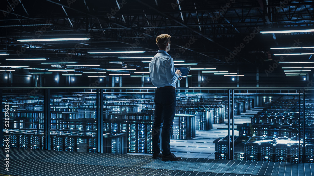 Poster Handsome Smiling IT Specialist Using Tablet Computer in Data Center. Succesful Businessman and e-Business Entrepreneur Overlooking Big Server Farm for Cloud Computing.