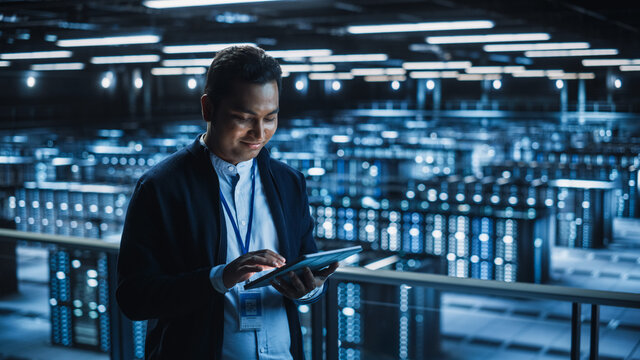 Handsome Smiling IT Specialist Using Tablet Computer in Data Center. Succesful Businessman and e-Business Entrepreneur Overlooking Server Farm Cloud Computing Facility. - Powered by Adobe