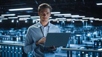 Handsome Smiling on Camera IT Specialist Using Laptop Computer in Data Center. Succesful Businessman and e-Business Entrepreneur Overlooking Server Farm Cloud Computing Facility.